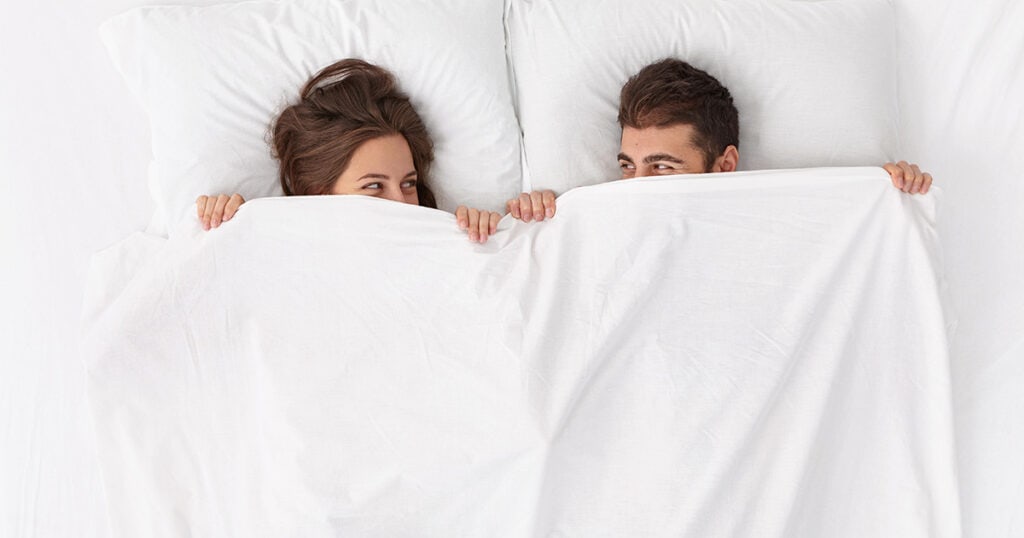 Couple in bed with sheets pulled up and looking at one another