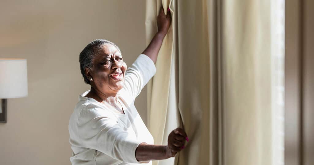 Older black woman open drapes, smiling and lit by sunshine